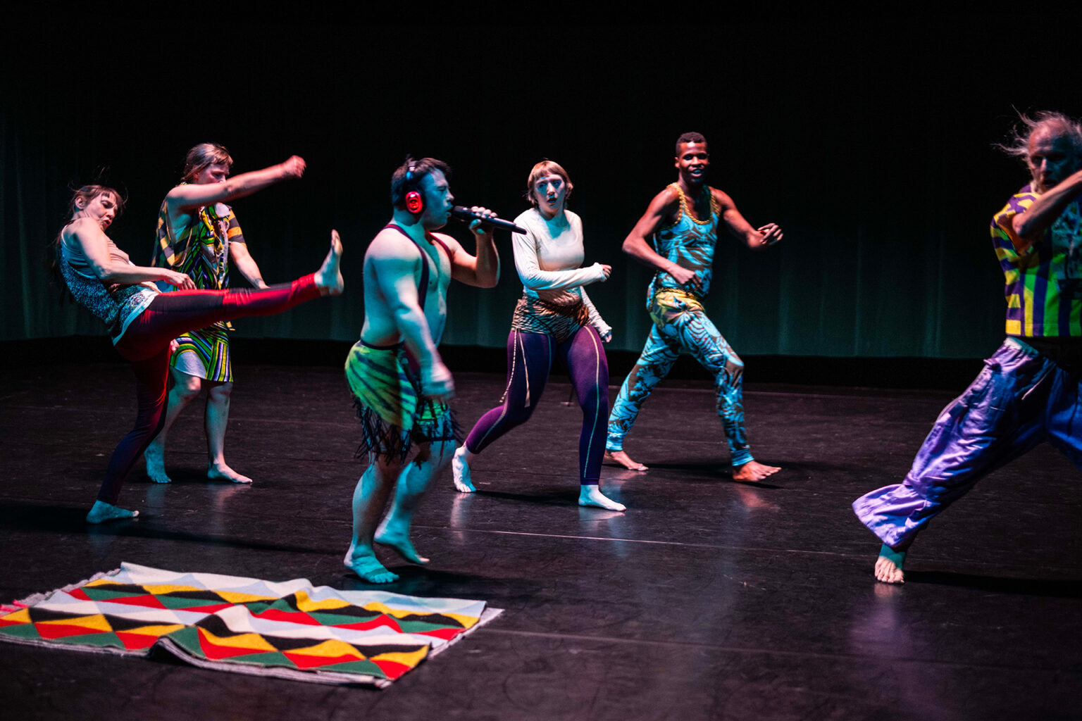 On voit les 6 danseurs, en vêtements bariolés, marcher vers la droite. L'un d'entre eux porte un micro à la main et chante.