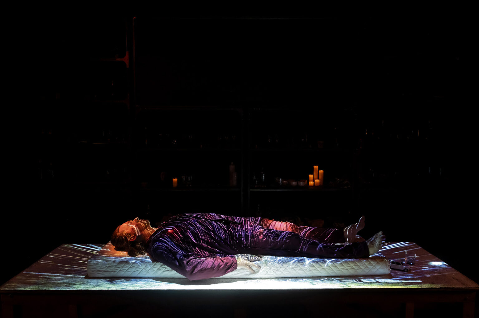 Le comédien Jarmo Reha est couché sur une grande table sur laquelle sont projetées des images de ville. Derrière lui, une étagère contient des verres et des bouteilles à foison.