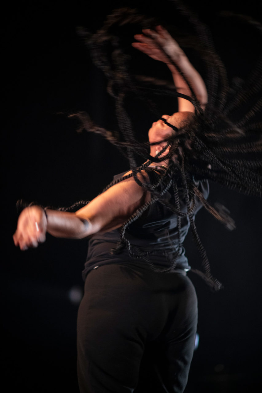 L'interprète Zena Moussa danse, de dos, les bras levés. Ses cheveux tressés volent dans tous les sens.