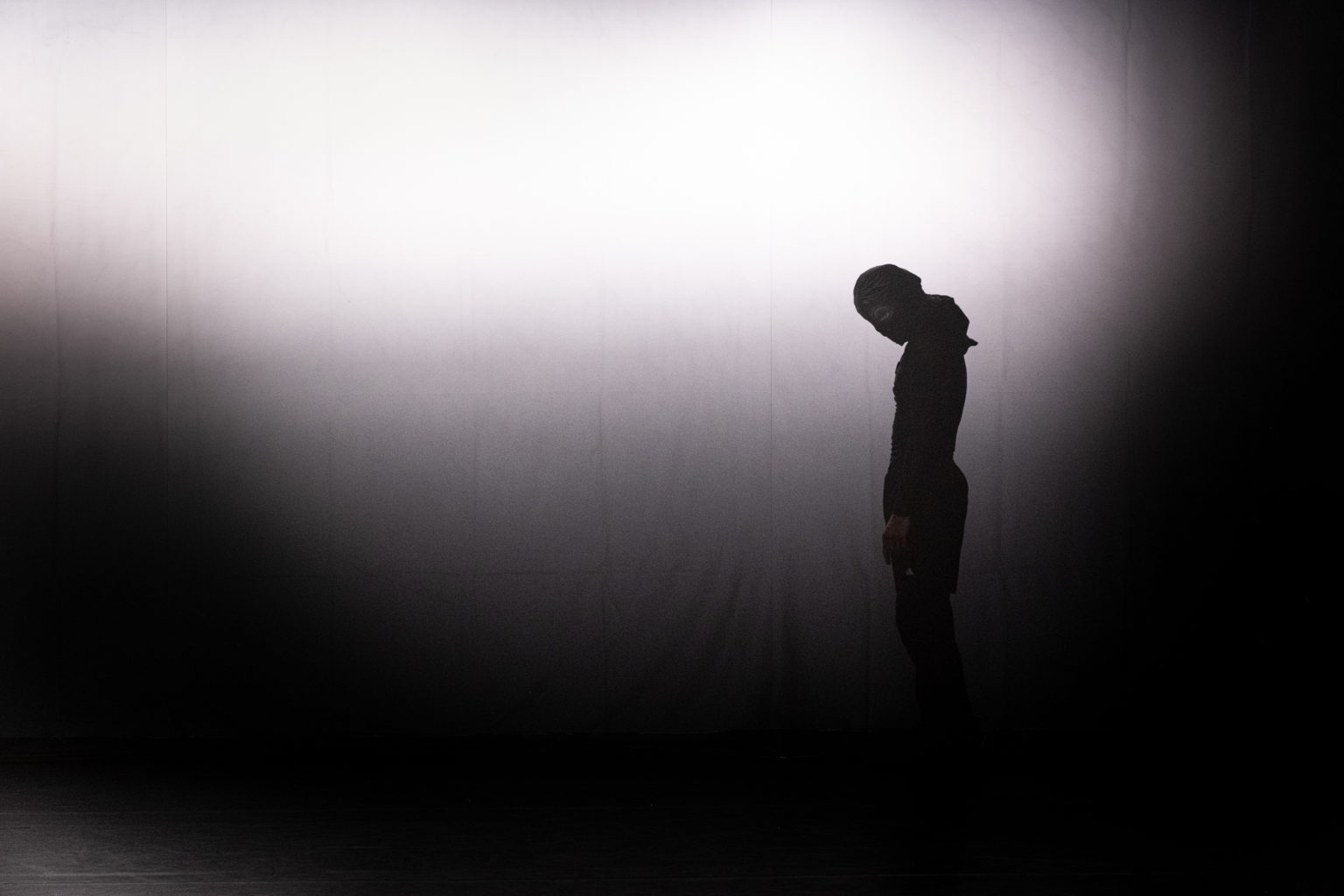 Mohamed Toukabri se tient debout, les mains le long du corps. Il est à contre-jour, une lumière forte se trouvant derrière lui. Il porte un masque.