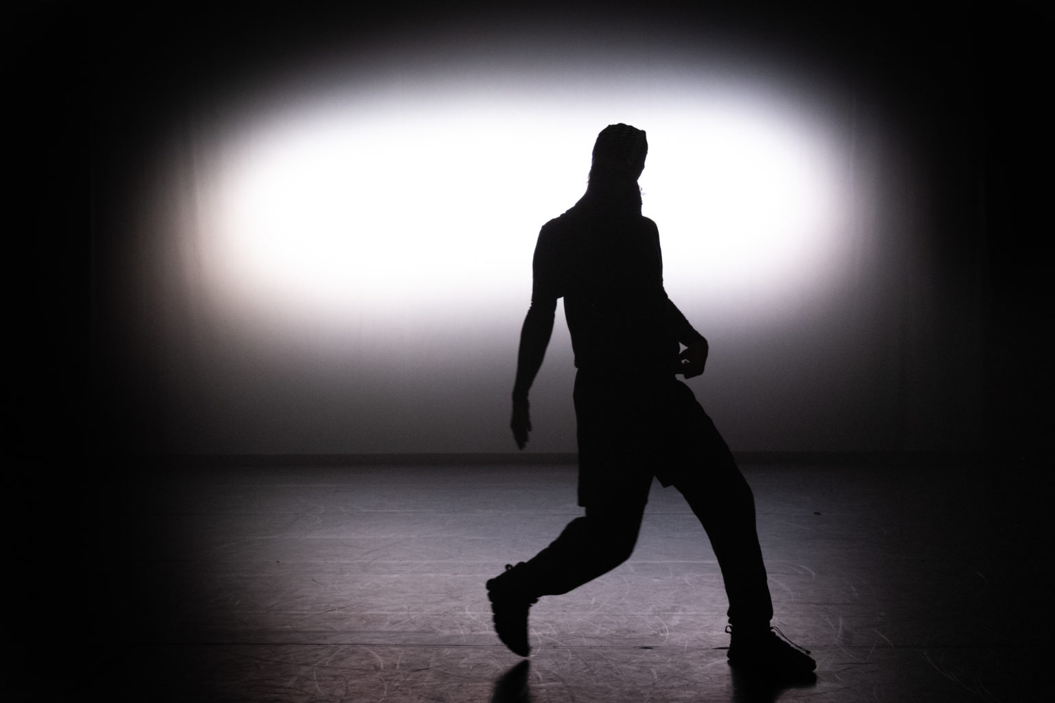 Mohamed Toukabri se tient debout, le corps en mouvement. Il est à contre-jour, une lumière forte se trouvant derrière lui.