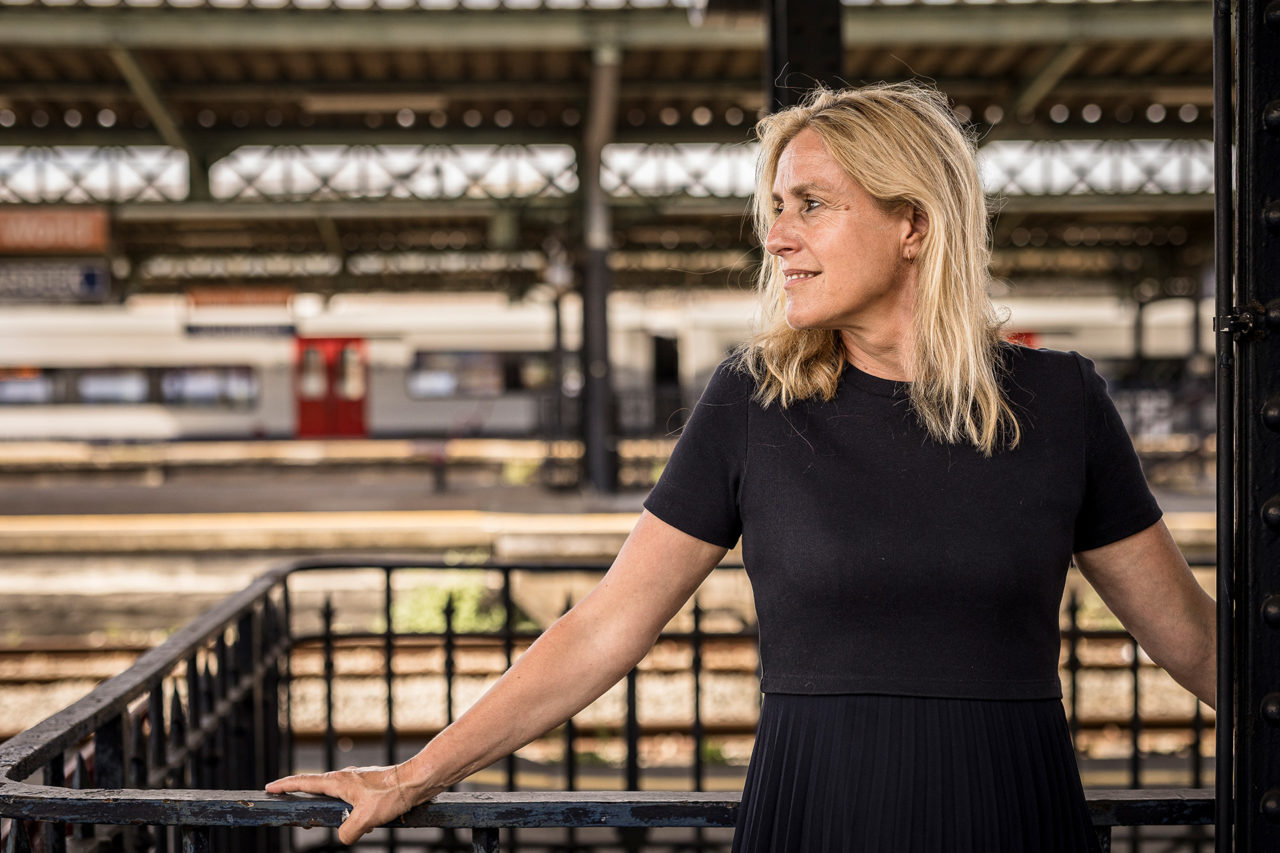 Geneviève Damas pose dans une gare. Elle est debout et se tient à une rambarde. Son visage est tourné vers la droite. Elle porte une robe noire.
