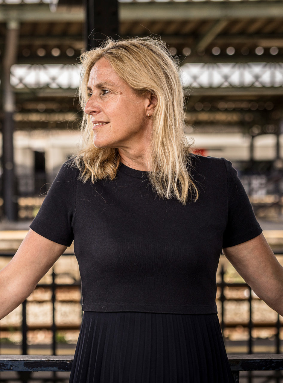 Geneviève Damas pose dans une gare. Elle est debout et se tient à une rambarde. Son visage est tourné vers la droite. Elle porte une robe noire.