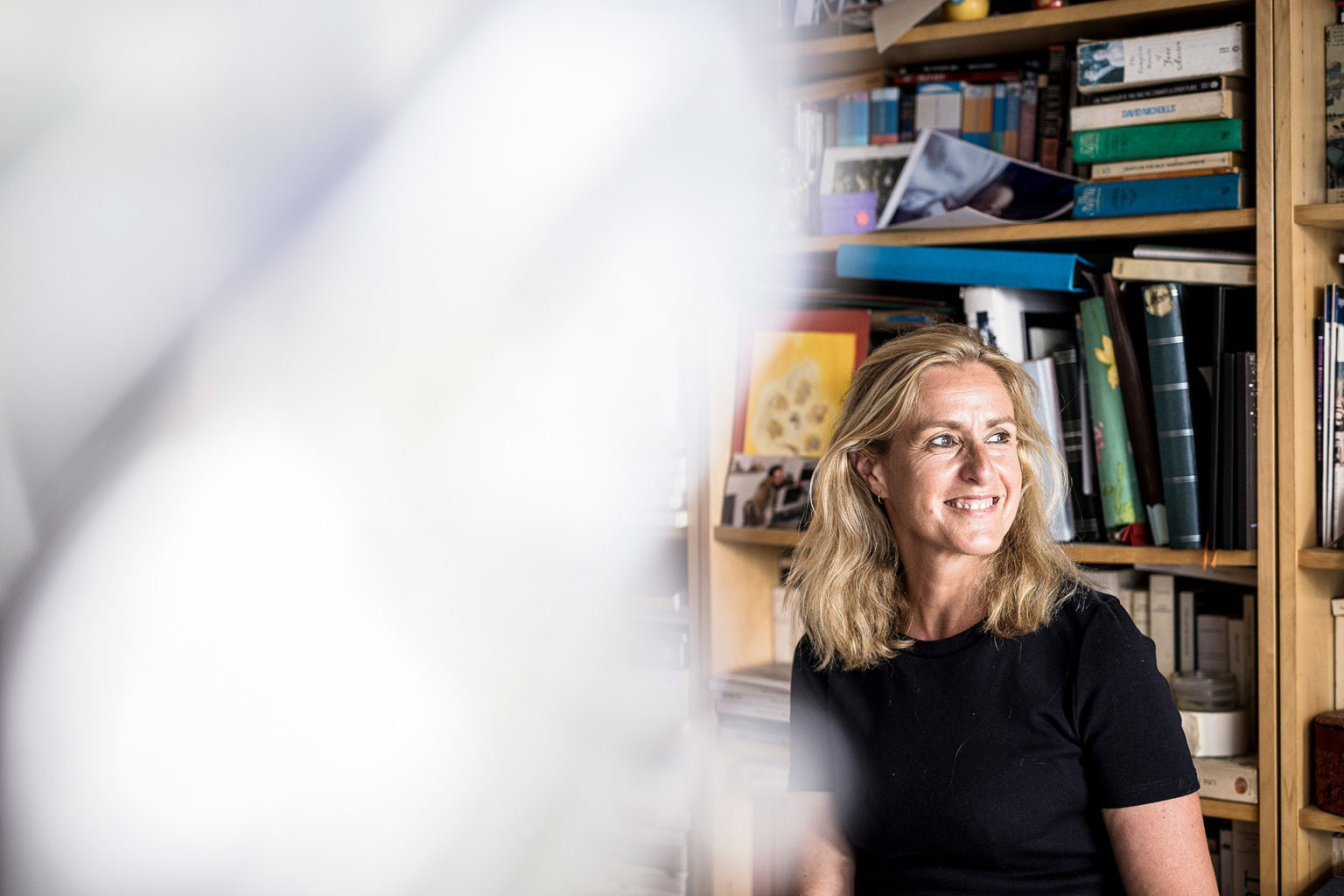 Geneviève Damas est assise devant une bibliothèque chargée de livres. Elle est souriante. Elle porte un haut noir.