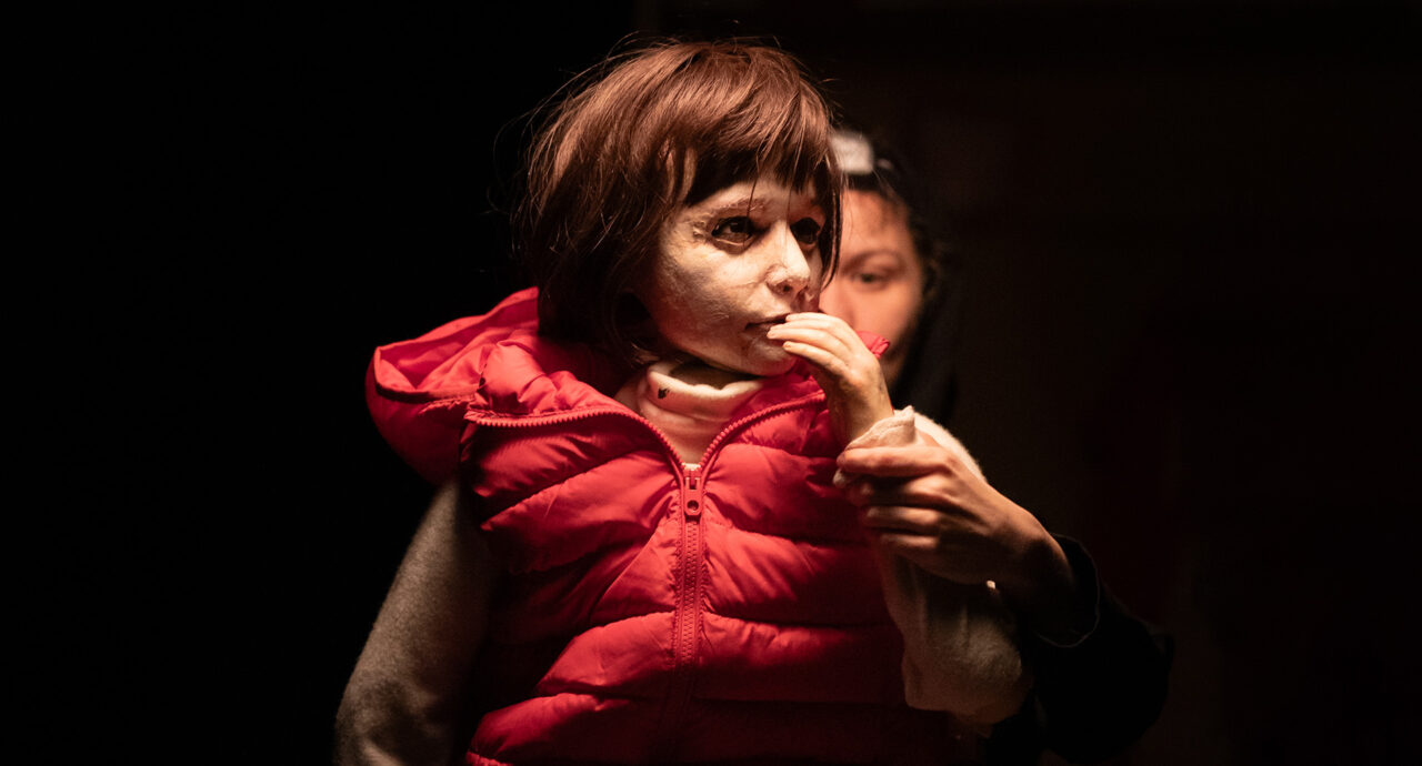 Une marionnette représentant une petite fille avec un gilet sans manches rouge. Au-dessus d'elle une lampe. Derrière elle, on devine la marionnettiste.