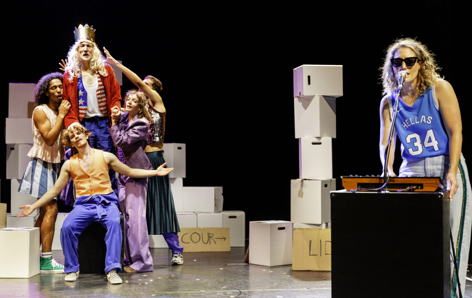 D'un côté de la scène (sur laquelle trônent plein de caisses), un groupe d'acteurs et actrices forment un bloc, comme une sculpture. Ils portent des vêtements colorés. De l'autre côté, il une femme qui porte des lunettes solaires et est devant une table de mixage.