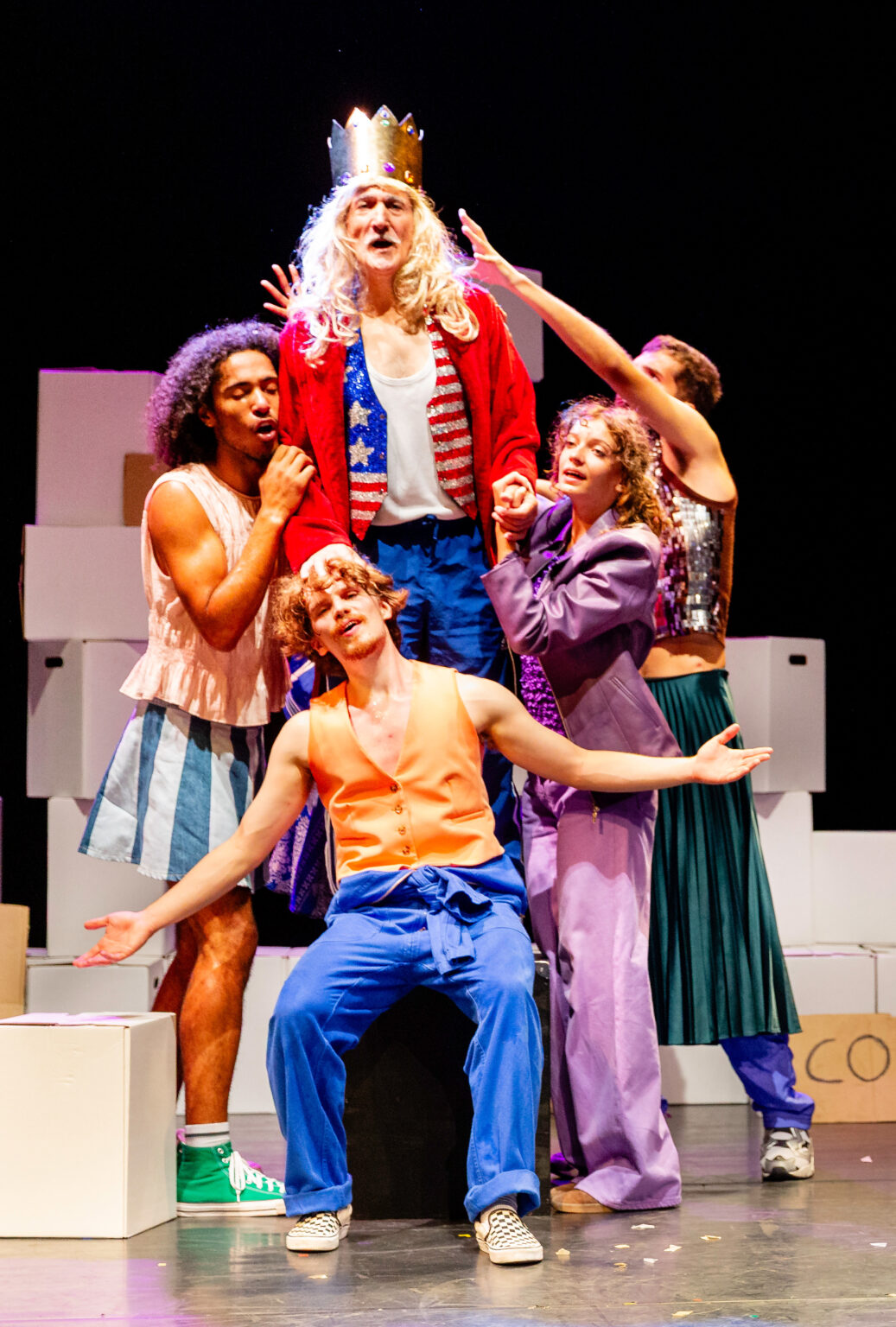 Sur une scène (sur laquelle trônent plein de caisses), un groupe d'acteurs et actrices forment un bloc, comme une sculpture. Ils portent des vêtements colorés.