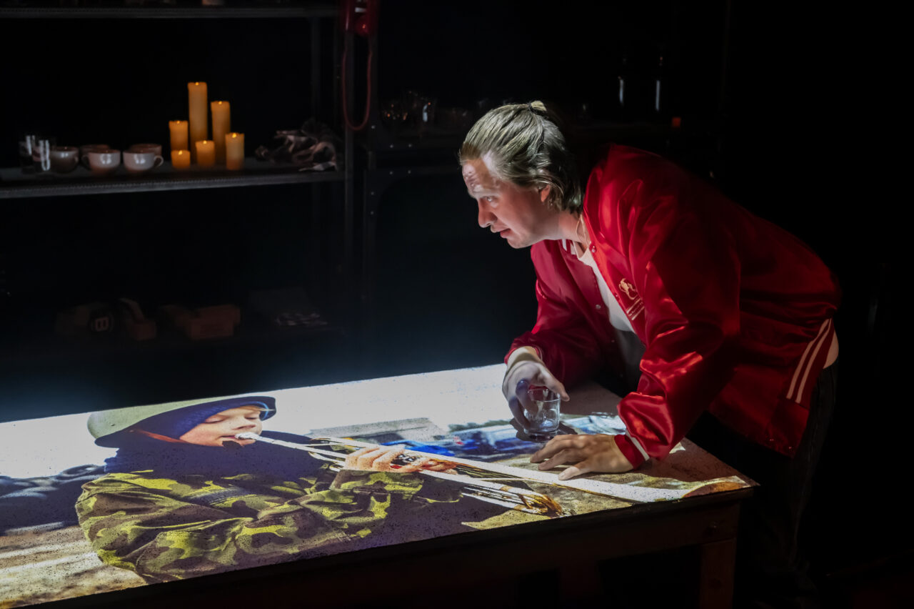 Un verre à la main, Jarmo Reha est penché sur une table sur laquelle est projetée l'image d'un petit garçon, habillé en militaire, qui fait de la trompette.