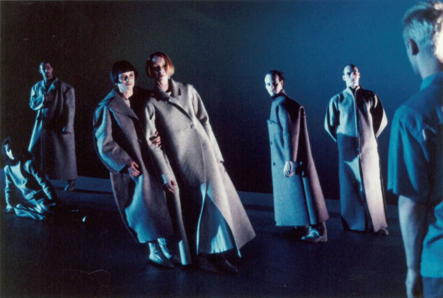 Sept personnes portant des longs manteaux d'hiver sont debout et regardent devant eux. Seul l'un d'entre eux a le dos tourné.