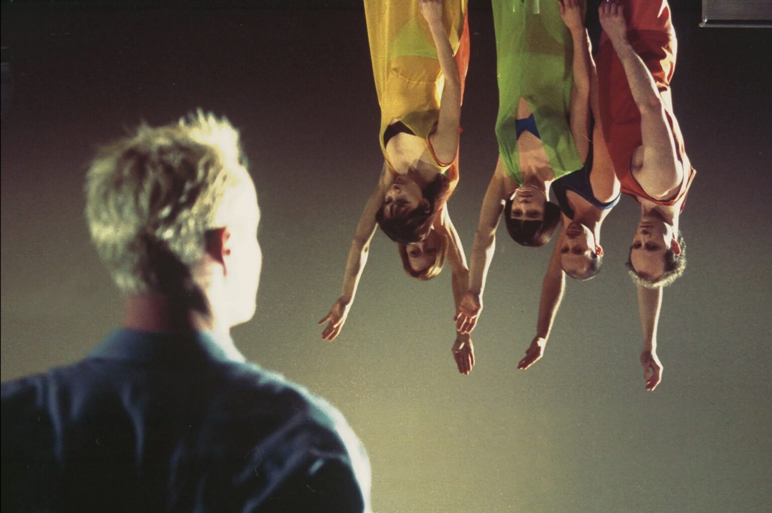 Un homme blond de dos à l'avant-plan regarde quatre personnes habillés de manière colorée, qui ont la tête en bas et lèvent le bras.