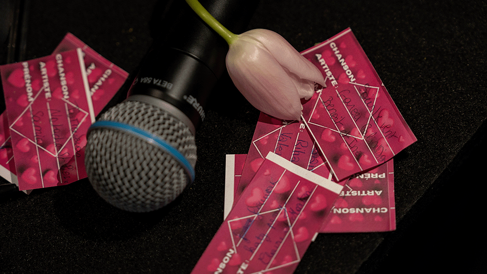 Sur un support noir, plusieurs cartes rouges comprenant le nom de chansons sont éparpillées. Par dessus les cartes, se trouvent un micro et une fleure rosée.