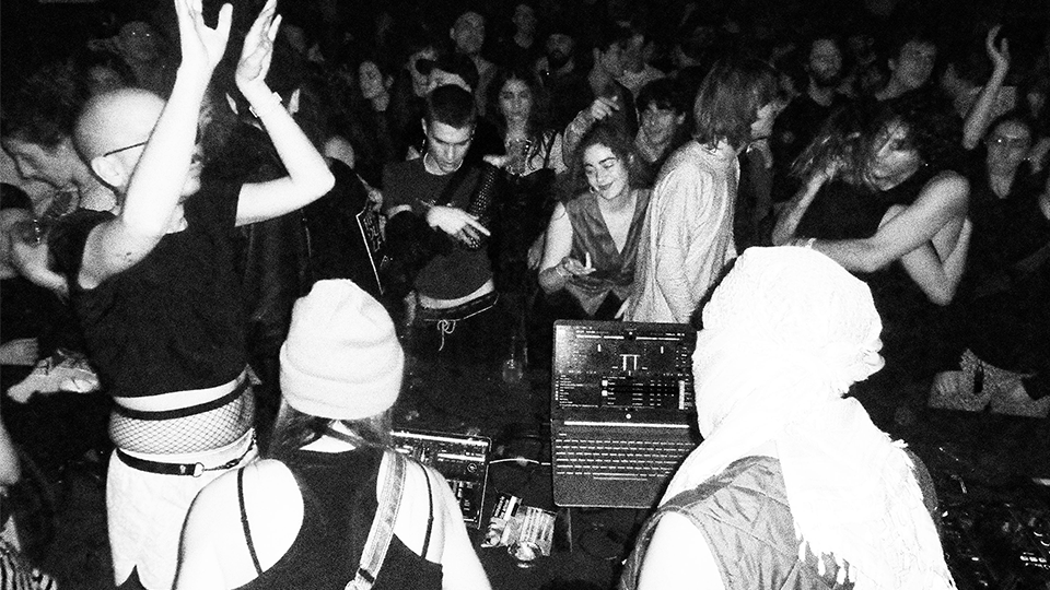 Photo en noir et blanc d'une foule en train de danser au milieu d'une salle sombre, deux personnes de dos sont face à des ordinateurs portables