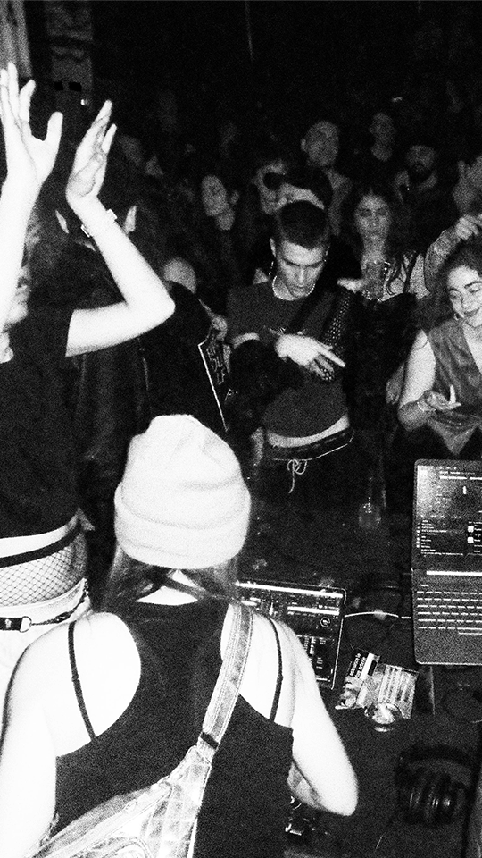Photo en noir et blanc d'une foule en train de danser au milieu d'une salle sombre, deux personnes de dos sont face à des ordinateurs portables