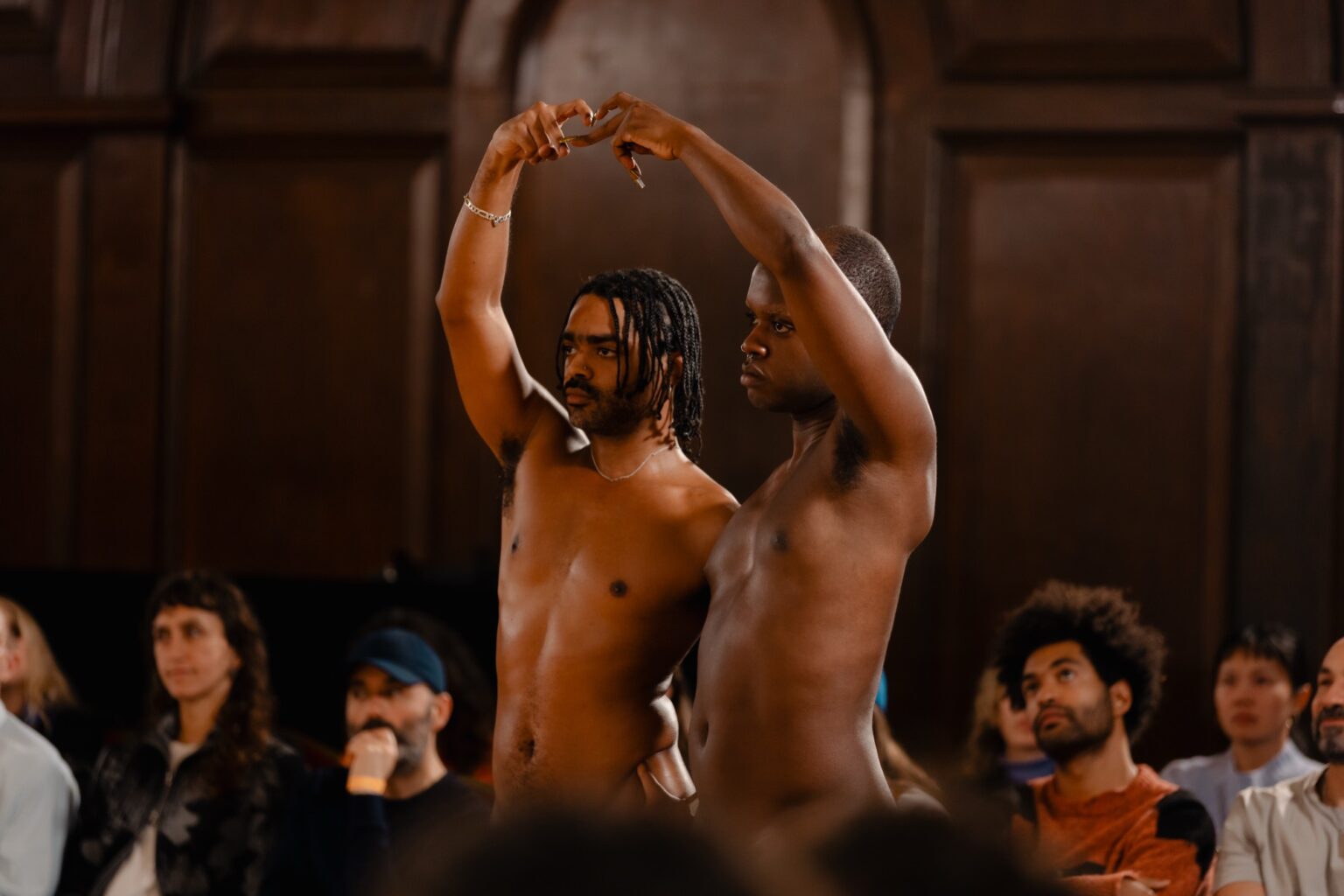 Deux personnes se tiennent debout nues, avec les bras formant un coeur. Derrière elles, on voit le public qui les regarde.