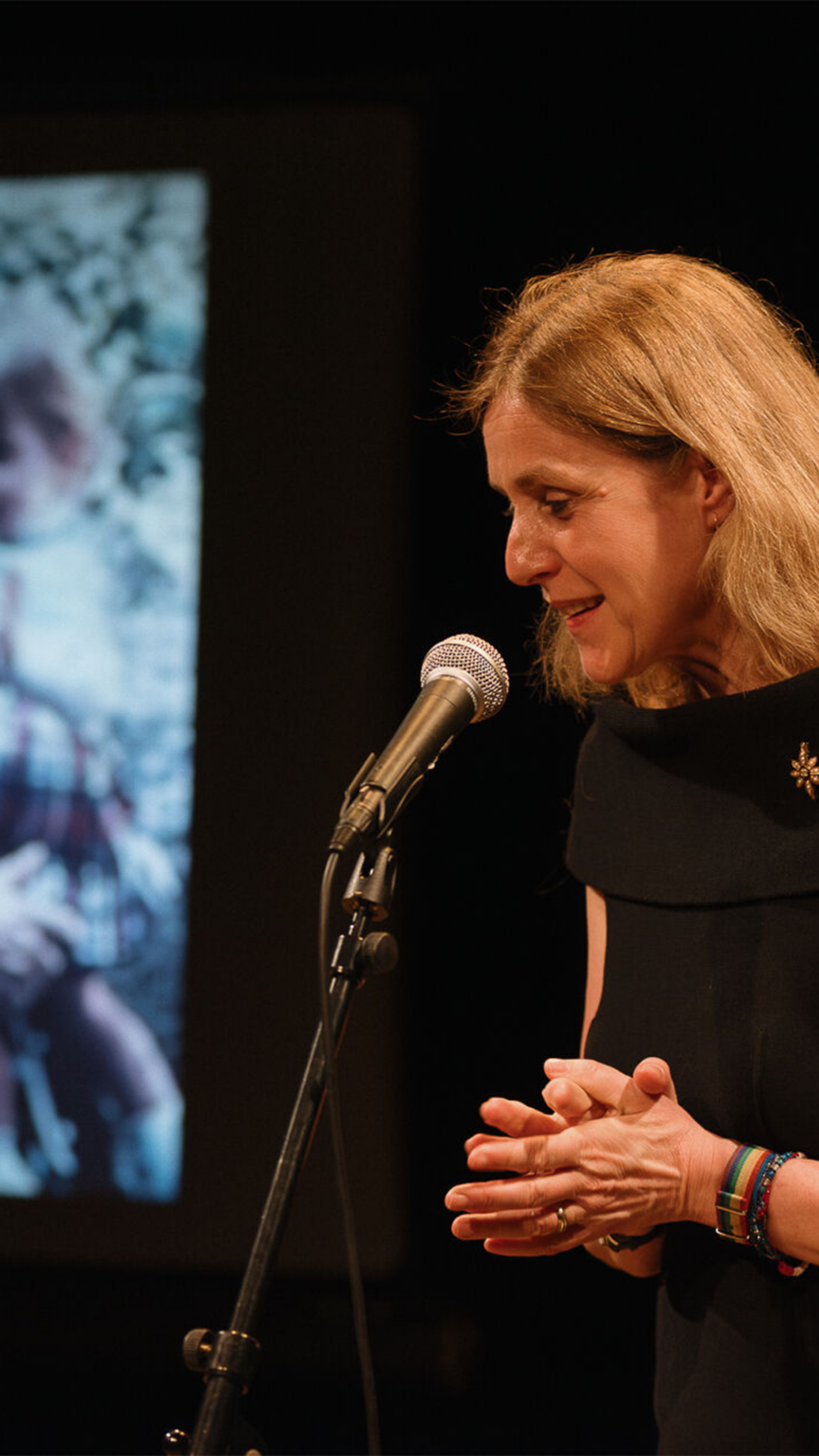 Geneviève Damas se tient debout, les mains jointes, derrière un micro. A coté d'elle, se trouve un écran montrant la photo d'une femme portant une petite fille.