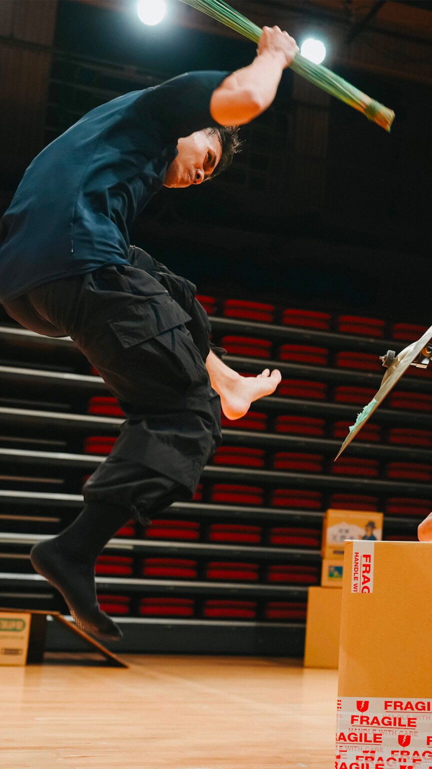 Un homme en scène saute en l'air, un pied levé plus haut que l'autre, une main portant une plante.
