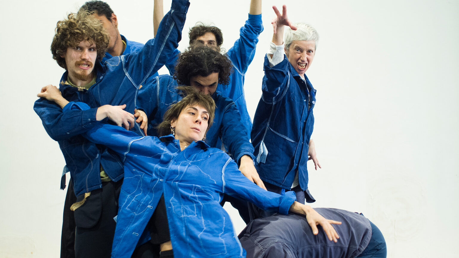 Groupe de plusieurs personnes en combinaisons bleues sur un fond clair. Une personne est soutenue par les autres. Des bras sont tendus dans différentes directions. Les corps sont rapprochés et partiellement superposés.
