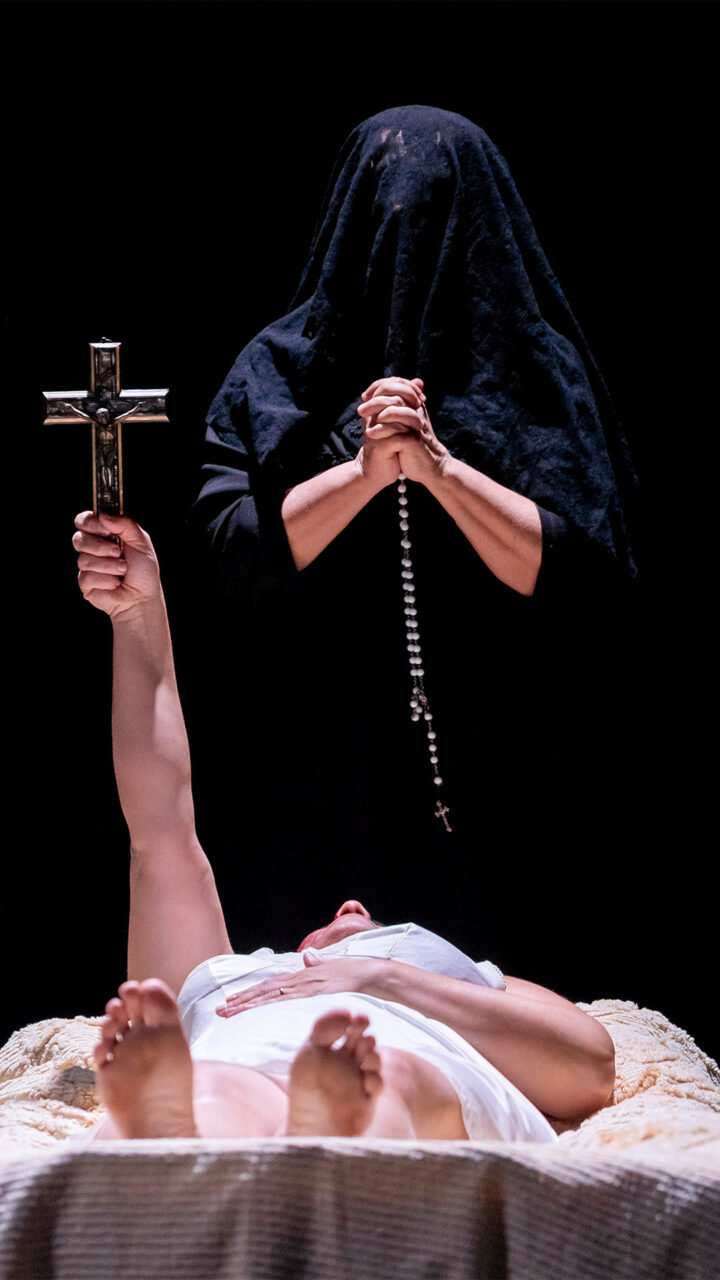 Une personne entièrement vêtue de noir se tient par dessus une autre personne allongée et recouverte d'un drap tenant une croix, le bras tendu vers le haut.