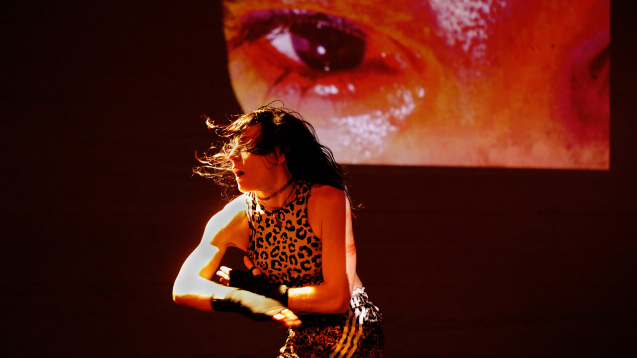 Marina Otero danse sur scène, derrière elle est projeté une image de visage rougeâtre. Elle porte des vêtements aux motifs de léopard.