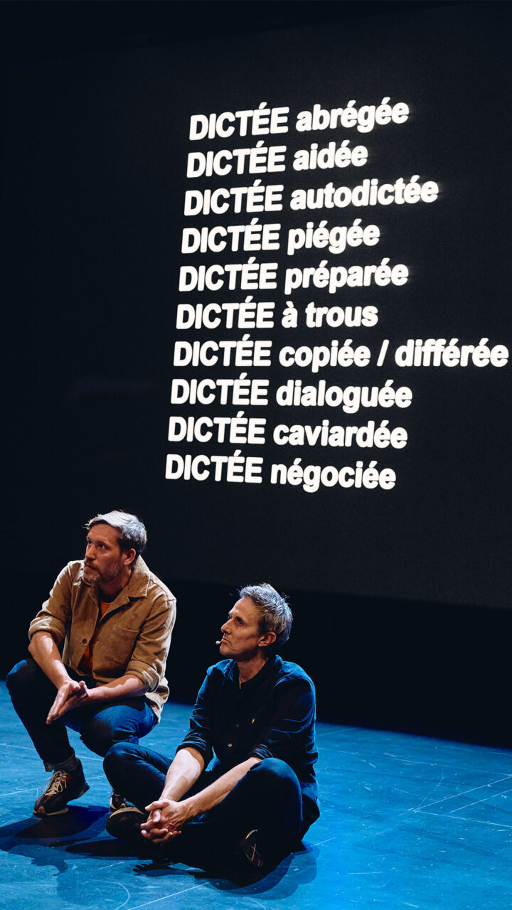 Arnaud Hoedt et Jérôme Prion sont assis sur scène. Derrière eux, sur un grand écran, apparaissent les mots d'une dictée, et plus précisément des participes passés.
