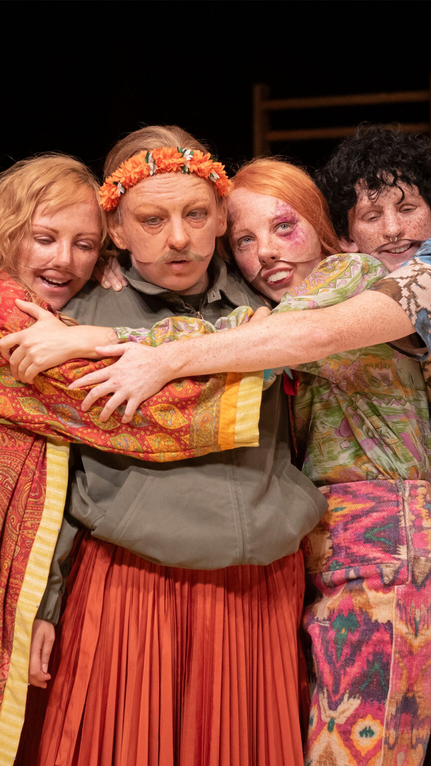 Photo du spectacle L'amour c'est pour du beurre où l'on voit 4 des comédiennes qui se font un câlin. Elles portent des masques réalistes et des vêtements colorés.