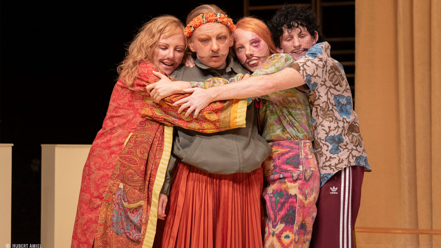 Photo du spectacle L'amour c'est pour du beurre où l'on voit 4 des comédiennes qui se font un câlin. Elles portent des masques réalistes et des vêtements colorés.