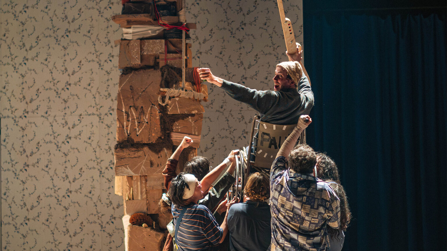 Scène de théâtre avec plusieurs personnes regroupées au pied d’une structure verticale faite de cartons et d’objets empilés. Une personne est en hauteur, appuyée sur l’installation. Des bras sont levés. Le décor comprend un mur clair à motifs et un rideau sombre sur le côté.