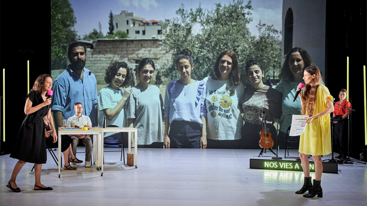 Scène de théâtre avec plusieurs intervenant·es sur un plateau. En fond, une grande projection montre un groupe de personnes en extérieur.