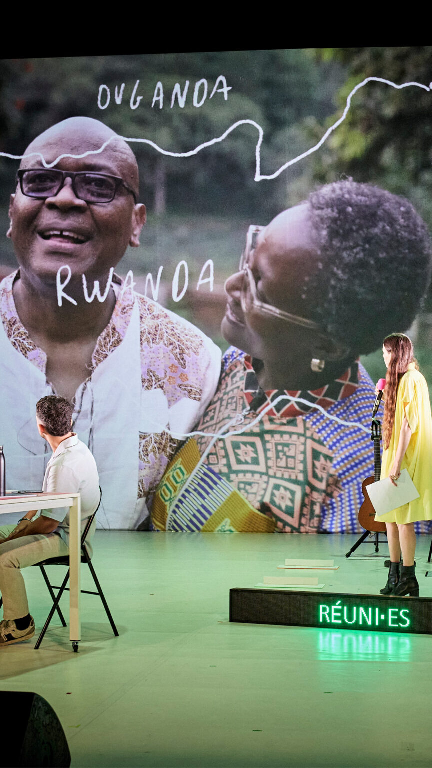 Plateau de théâtre avec plusieurs personnes réparties dans l’espace. En fond, une grande image projetée montre deux personnes devant un décor naturel. Des mots sont dessinés sur l’image, dont « RWANDA ». Des tables, des micros et un écran lumineux au sol sont visibles.