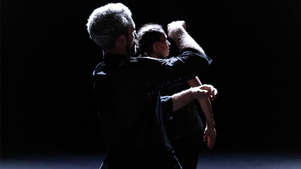Micael se tient dos à la caméra devant Angela, se bras droit forme un angle droit vers le haut. Le fond est obscur mais le sol est blanc