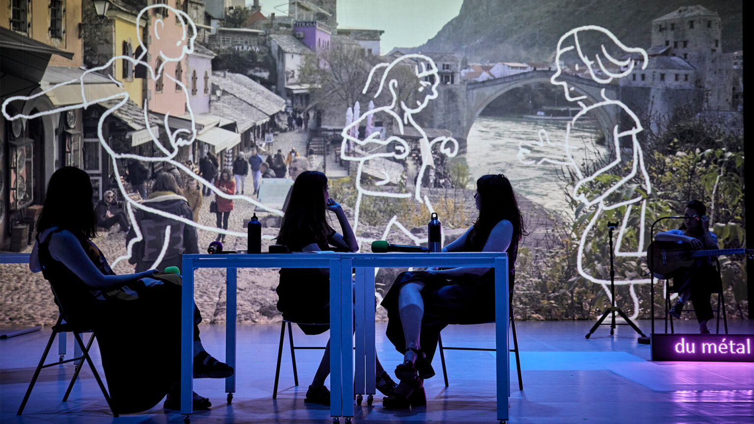 Les trois interprètes du spectacle - Oksana Leuta (qui vient d'Ukraine), Hala Rajab (qui vient de Syrie), Ines Tanović (qui vient de Bosnie) - sont autour d'une table. Elles sont dans la pénombre et regardent le grand écran derrière elles sur lequel est projeté une photo du pont de Mostar et des dessins d'enfants en train de jouer par-dessus la photo.