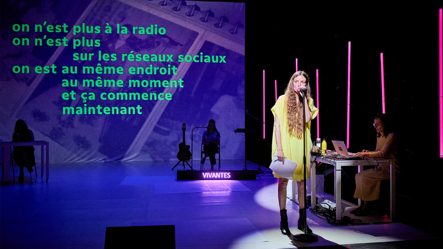 Aurélie Charon est à l'avant-scène et parle dans un micro sur pied. Elle porte une robe jaune. Derrière elle, le reste de la scène est dans la pénombre. Des personnes sont assises dans le fond et du texte apparait sur un grand écran dans le fond de la scène.