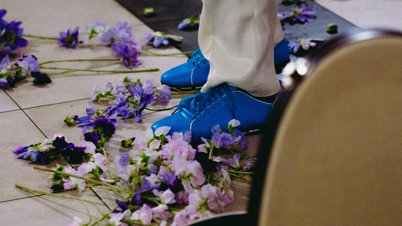 Des chaussures bleues sont visibles au sol. Le sol est recouvert de fleurs violettes et roses. Le bas d’un vêtement clair est visible. Les fleurs sont éparpillées autour des pieds. Une chaise apparaît partiellement sur le côté.