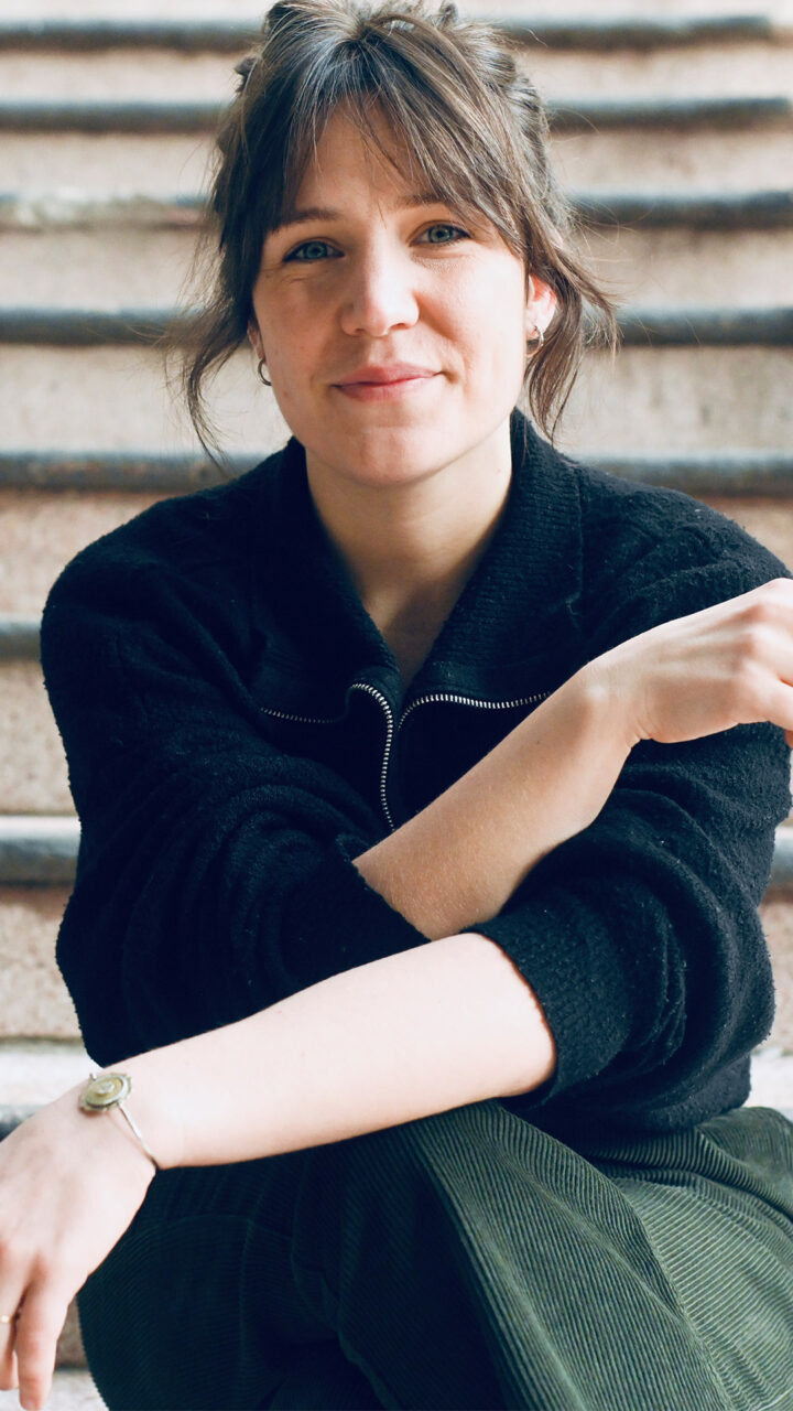 Portrait d’Héloïse Ravet assise sur un escalier. Le regard est dirigé vers l’objectif. Les mains sont visibles au premier plan. L’escalier forme des lignes horizontales en arrière-plan. Un garde-corps métallique est visible sur le côté.