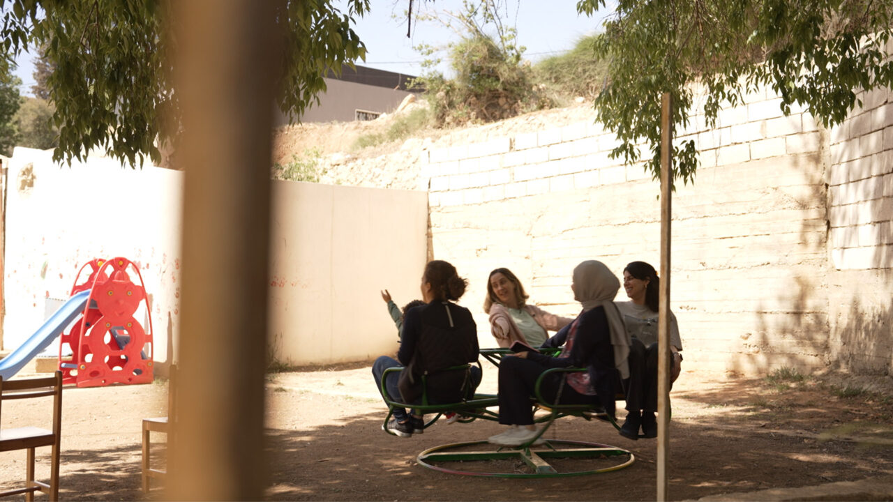 Quatre personnes sont assises sur un manège circulaire dans un espace extérieur. Des arbres projettent de l’ombre. Un mur entoure le lieu. Une structure de jeu est visible à gauche.