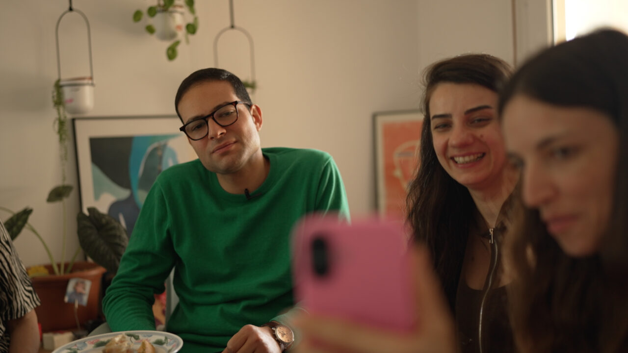 Trois personnes sont assises autour d’une table dans un intérieur. Une personne tient un téléphone au premier plan. Une assiette et des objets décoratifs sont visibles.