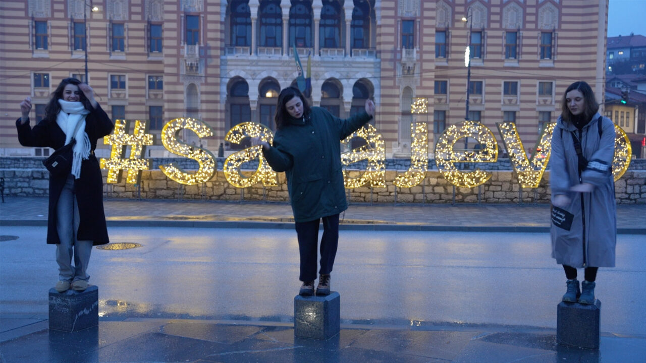 Trois personnes se tiennent sur des plots dans une rue. Des lettres lumineuses forment le mot "Sarajevo" en arrière-plan. Un bâtiment avec une façade décorée est visible derrière.