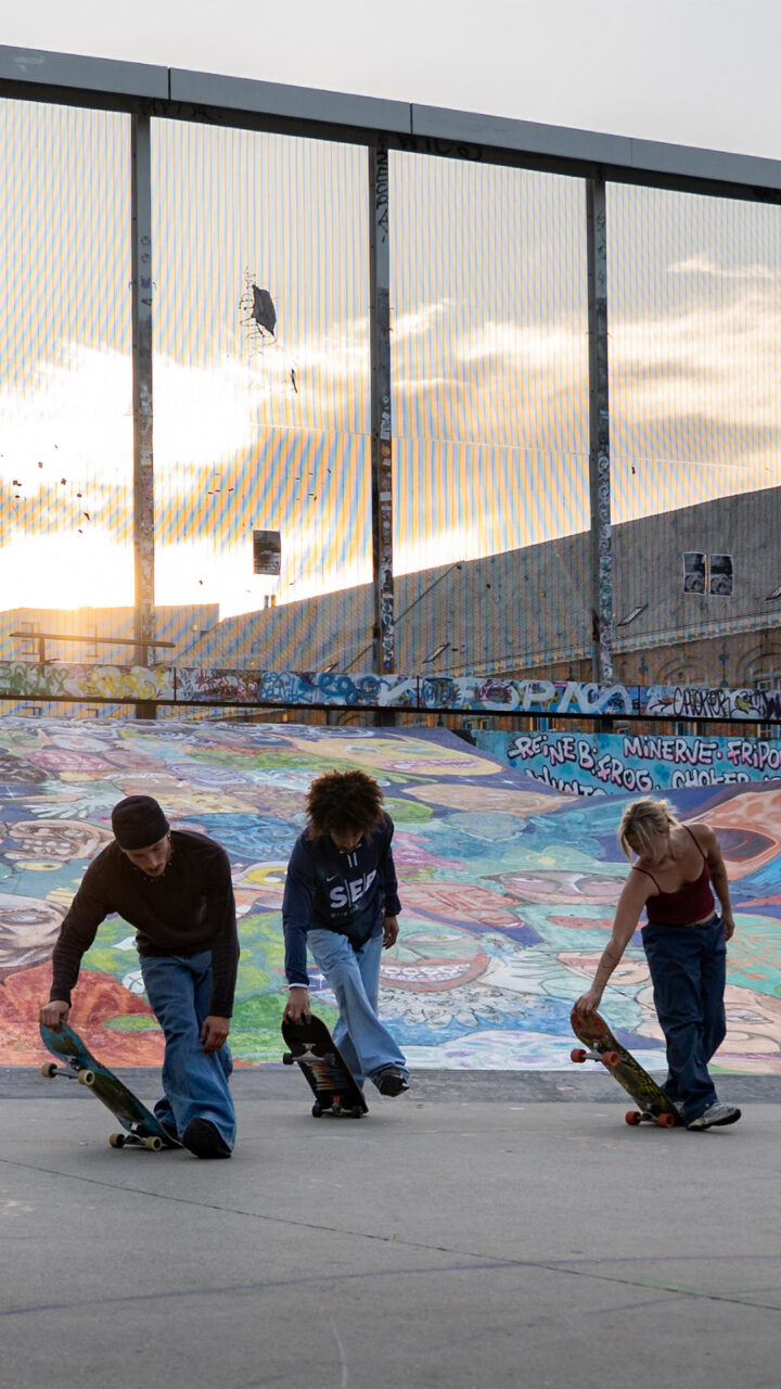 5 danseurs et skateurs sont penchés et tiennent un skateboard en mains. Le soleil décline au loin. La scène se passe au Skatepark des Ursulines.