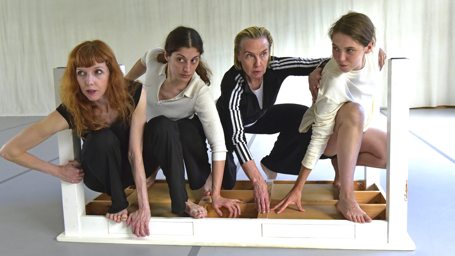 Les 4 interprètes de Lignes de fracture (Caterina Campo, Michèle Noiret, Lise Vachon, Léa Vinette) sont accroupies les unes à côté des autres et se tiennent dans une table blanche retournée.