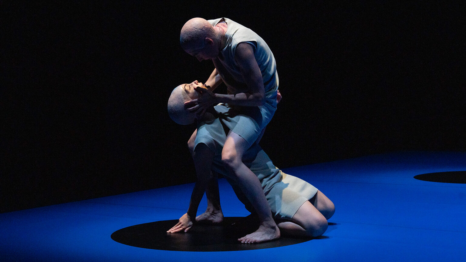 Un danseur tient dans ses mains le visage d'une danseuse, accroupie. Il la maintient à terre, entre ses jambes. Tous deux portent tous une robe blanche.