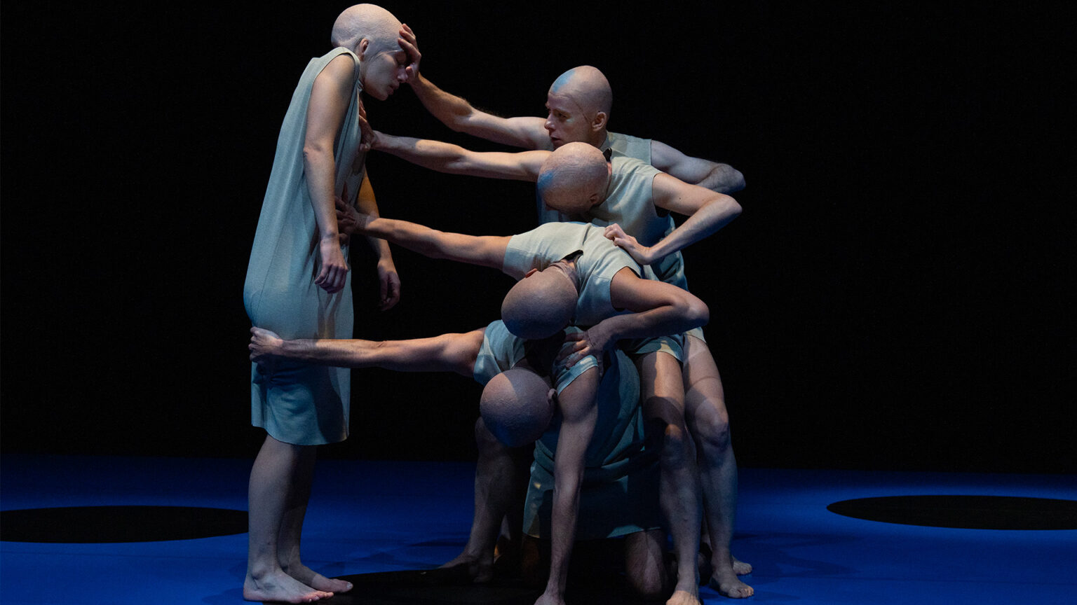 Une danseuse est debout. A côté d'elle, quatre danseurs se superposent les uns aux autres et tentent de la repousser avec leurs bras. Ils portent tous une robe blanche.