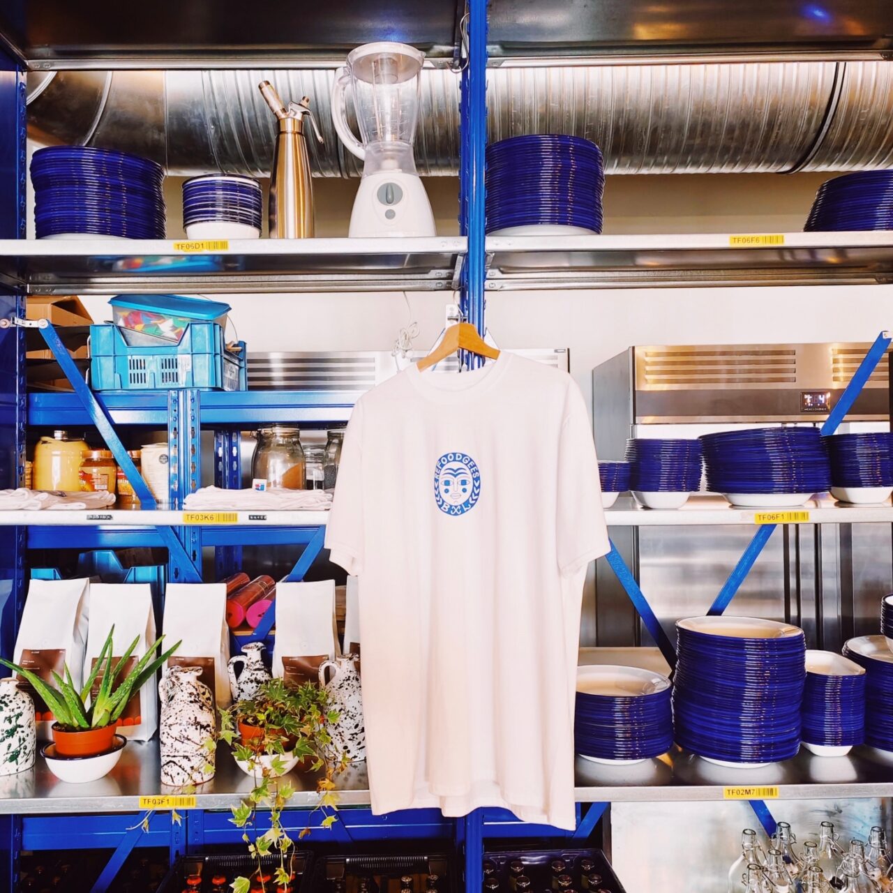 On voit des étagères bleues avec de la vaisselle bleue et blanche, un t-shirt avec le logo de Refoodgees, des plantes et différents ustensiles de cuisine.