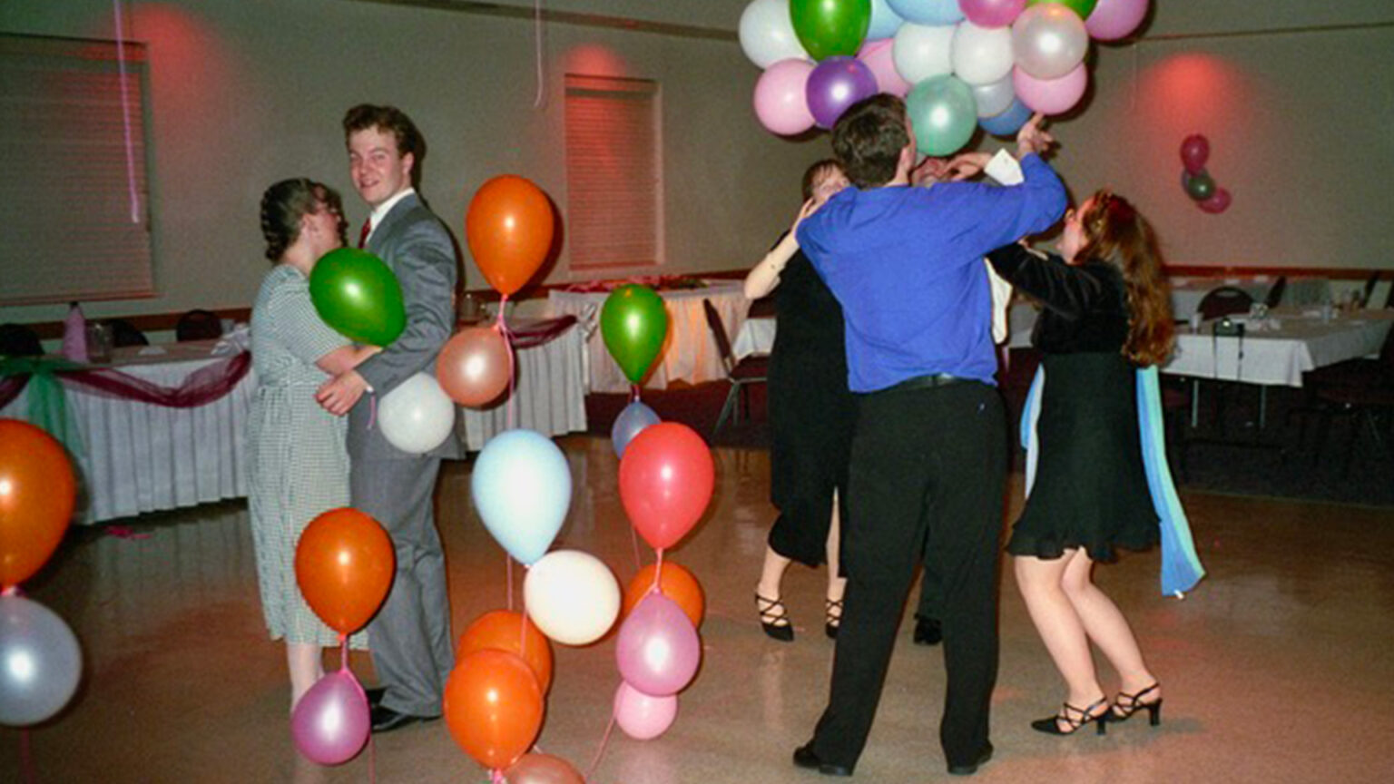 Des personnes dansent à une fête de famille, dans une salle de fête. Il y a des ballons de toutes les couleurs.